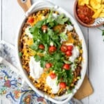A casserole dish filled with a baked Mexican-inspired dish topped with melted cheese, sour cream, cherry tomatoes, cilantro, and green onions—perfect for june 30 minute dinners. A bowl of salsa and chips is on the side.