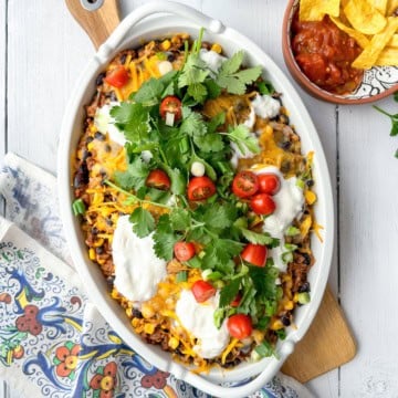 A casserole dish filled with a baked Mexican-inspired dish topped with melted cheese, sour cream, cherry tomatoes, cilantro, and green onions-perfect for june 30 minute dinners. A bowl of salsa and chips is on the side.