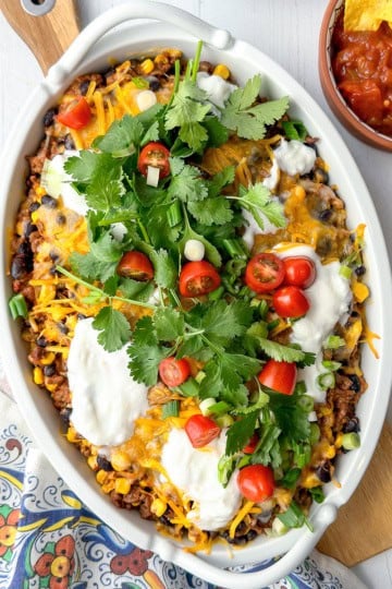 A casserole dish filled with a baked Mexican-inspired dish topped with melted cheese, sour cream, cherry tomatoes, cilantro, and green onions—perfect for june 30 minute dinners. A bowl of salsa and chips is on the side.