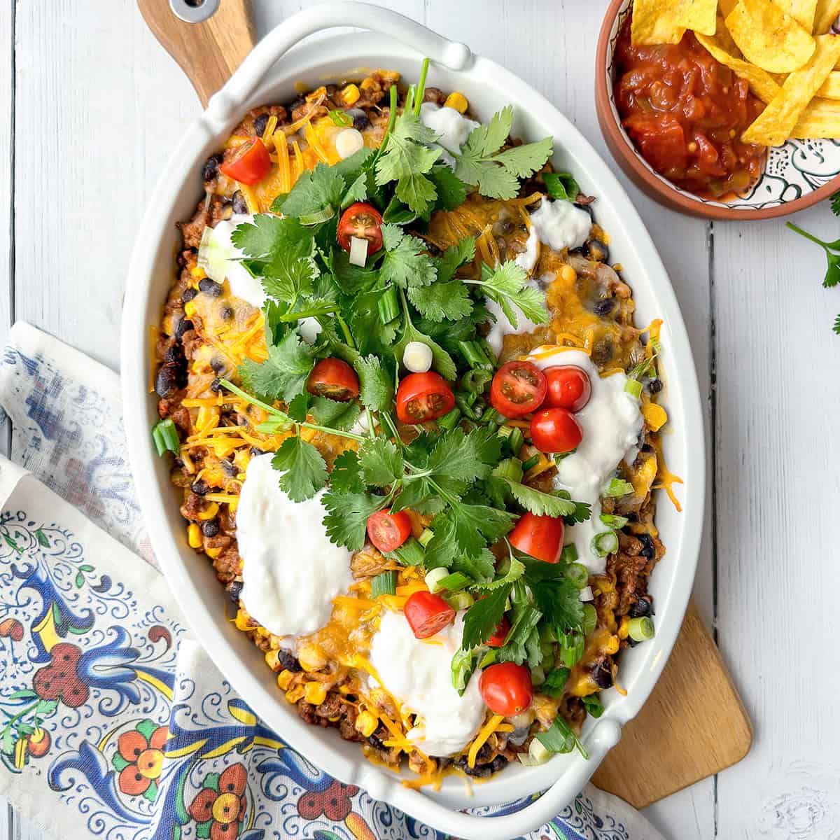 A casserole dish filled with a baked Mexican-inspired dish topped with melted cheese, sour cream, cherry tomatoes, cilantro, and green onions-perfect for june 30 minute dinners. A bowl of salsa and chips is on the side.