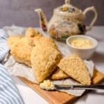 A batch of colonial scones on a wooden board with a knife, a dish of clotted cream, and a decorative teapot in the background—perfect for an inviting tea table setting.