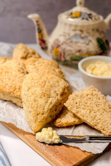 A batch of colonial scones on a wooden board with a knife, a dish of clotted cream, and a decorative teapot in the background—perfect for an inviting tea table setting.