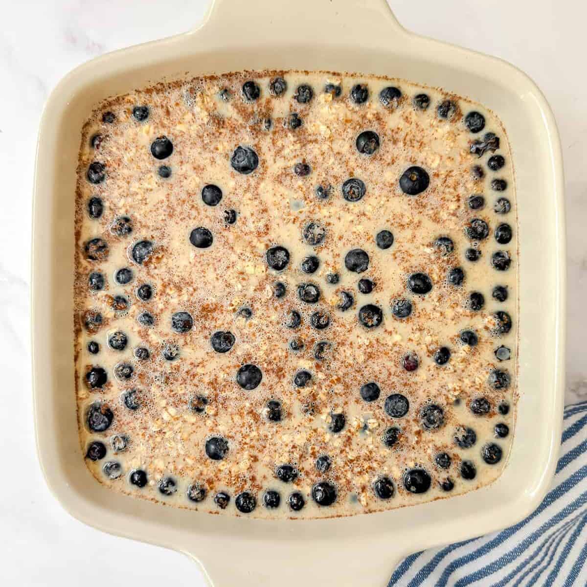 A square baking dish filled with unbaked blueberry Amish baked oatmeal batter sits on a white surface next to a blue and white striped cloth.