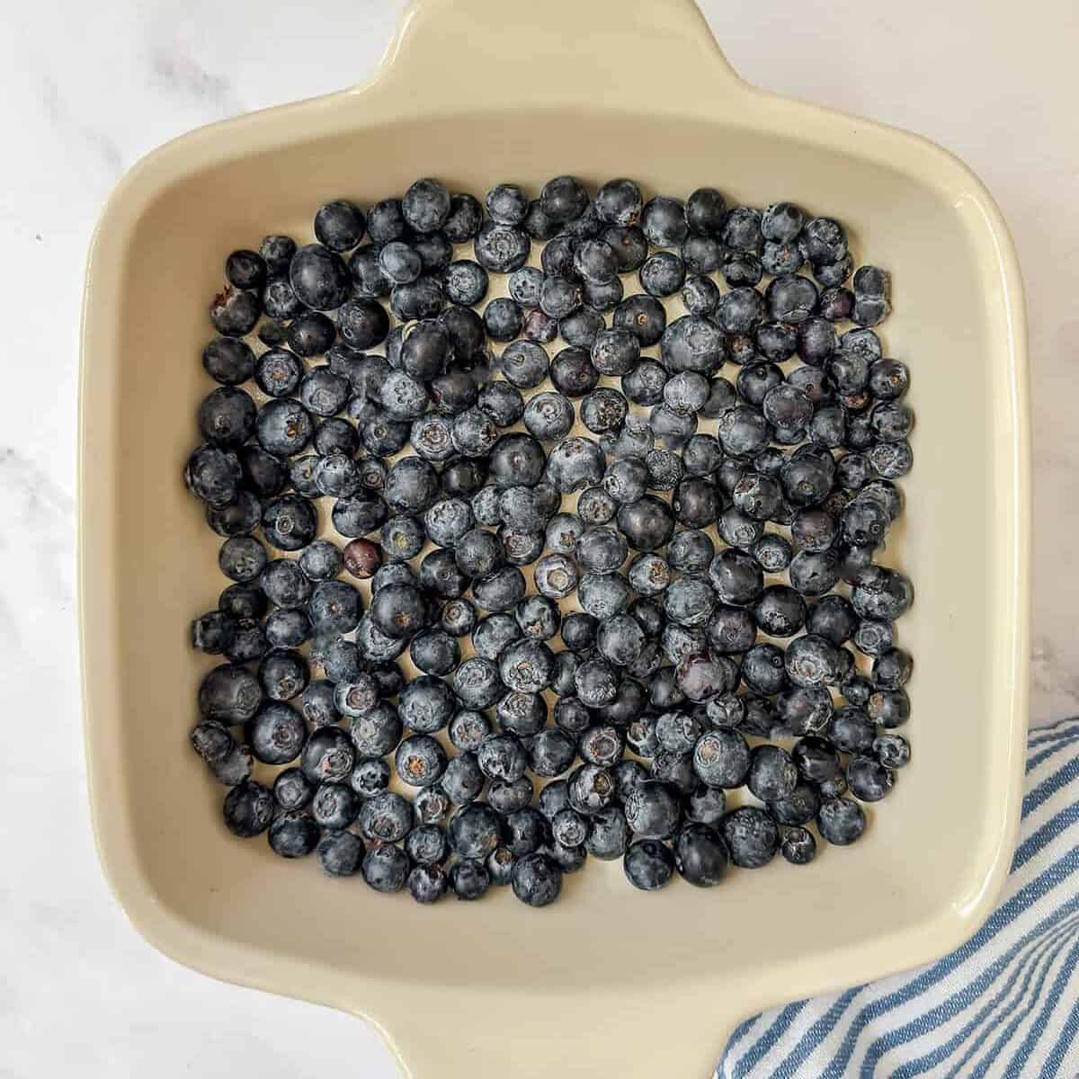 A square ceramic baking dish filled with a single layer of fresh blueberries, perfect for blueberry Amish baked oatmeal, sits next to a blue and white striped cloth on a white surface.