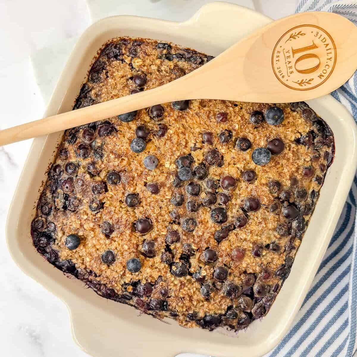A blueberry Amish baked oatmeal dish sits in a square white baking pan, topped with juicy blueberries, with a wooden spoon resting on top and a bowl of fresh blueberries nearby.