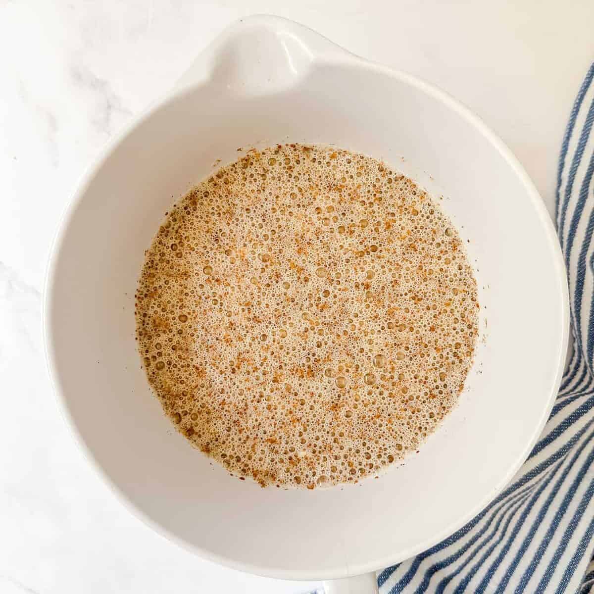 A white mixing bowl containing a frothy brown liquid mixture for blueberry Amish baked oatmeal, placed on a white surface next to a blue and white striped cloth.