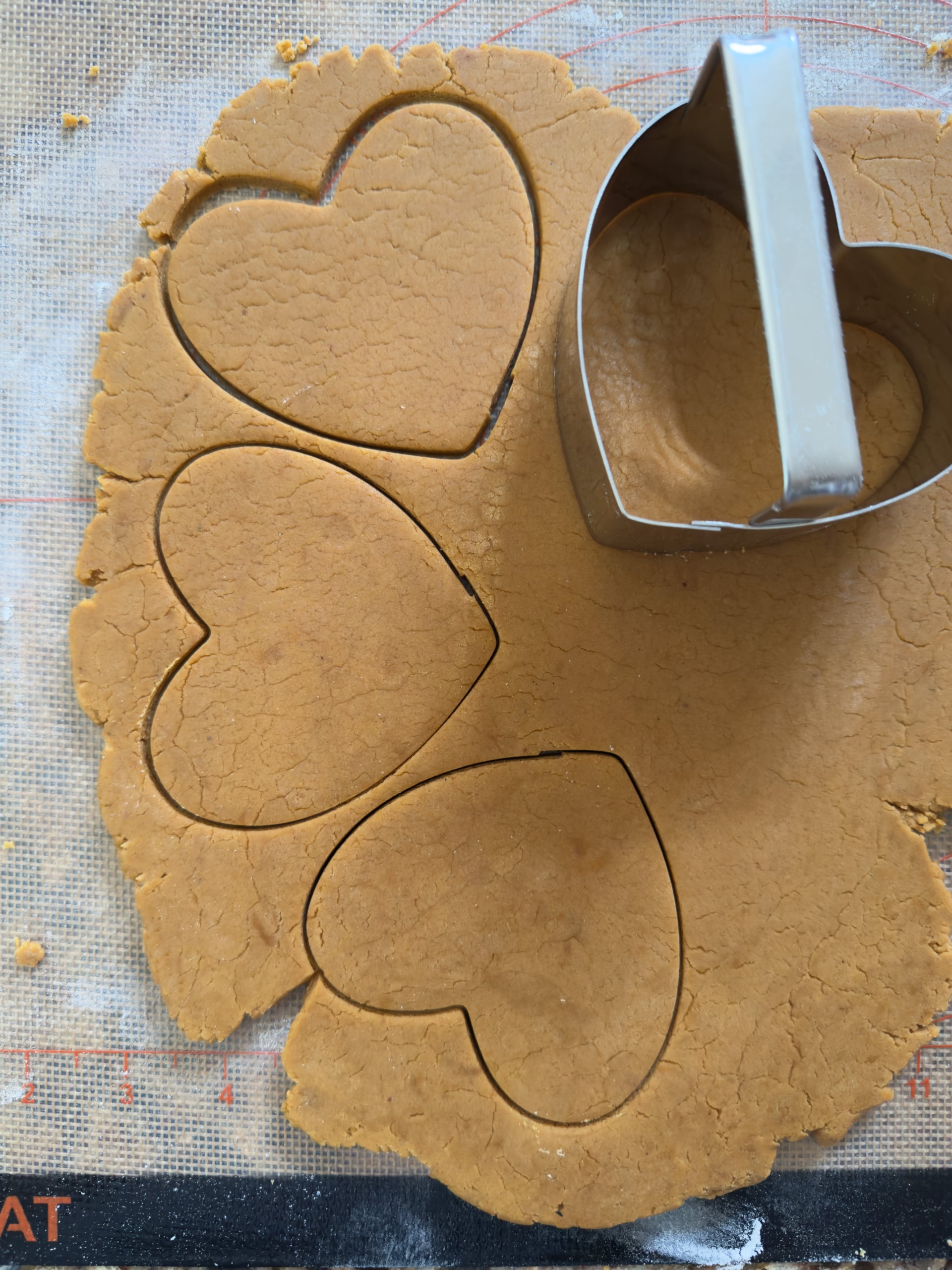 A metal heart-shaped cookie cutter cuts heart shapes from rolled dough for gingerbread cookies on a silicone baking mat.
