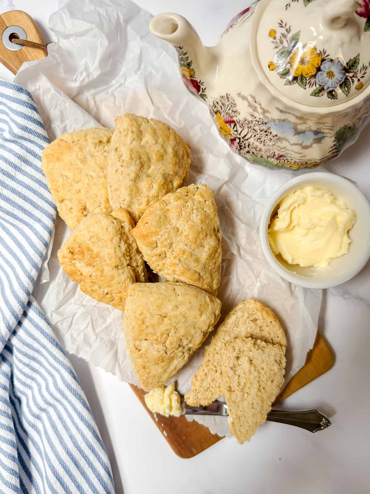 Six Colonial Scones are arranged on parchment paper with one split open, served with a bowl of butter and a floral teapot—perfect for enjoying at a Tea Table on a bright white surface.