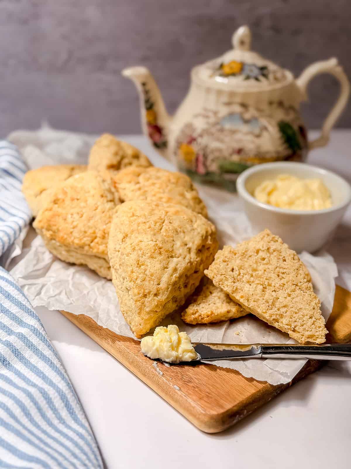Triangular Colonial Scones on parchment paper atop a wooden board, with a knife holding clotted cream, a bowl of cream, and a decorative teapot evoke the charm of an Early American Recipe perfect for any tea table.