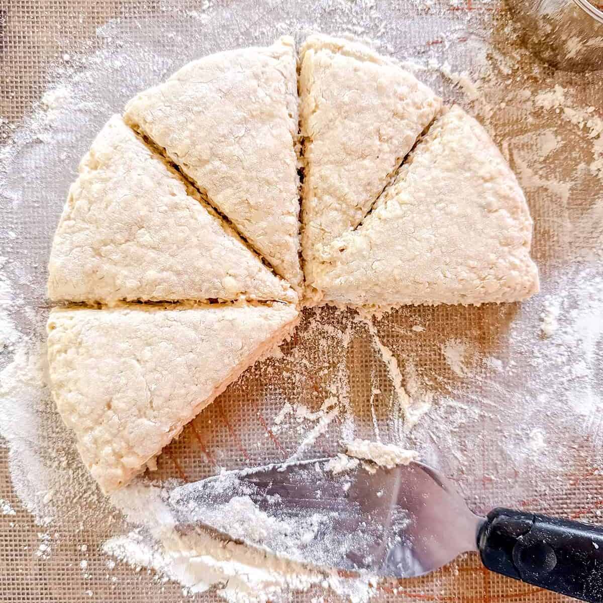 A round dough cut into six wedges sits on a floured surface, ready to become Colonial scones-a nod to early American tea table traditions-with a metal spatula and biscuit cutter nearby.
