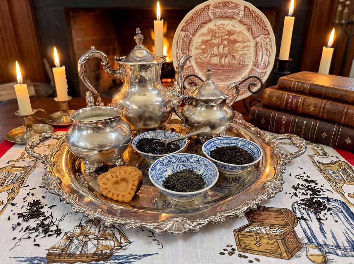 Silver colonial-era tea service with three blue and white Chinese tea bowls displaying loose leaf tea varieties from the Boston Tea Party manifest, set on a nautical tablecloth beside lit candles and a glowing fireplace, with Tea Act and Common Sense books visible in the background.