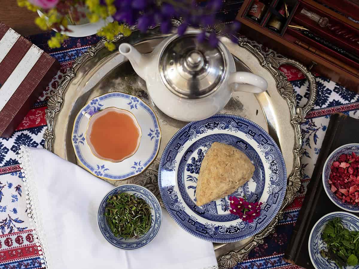 A silver tray holds a teapot, a cup of Liberty Tea recipe, a blue-patterned plate with a scone, small bowls with herbs and dried berries, and a white napkin on a patterned tablecloth.
