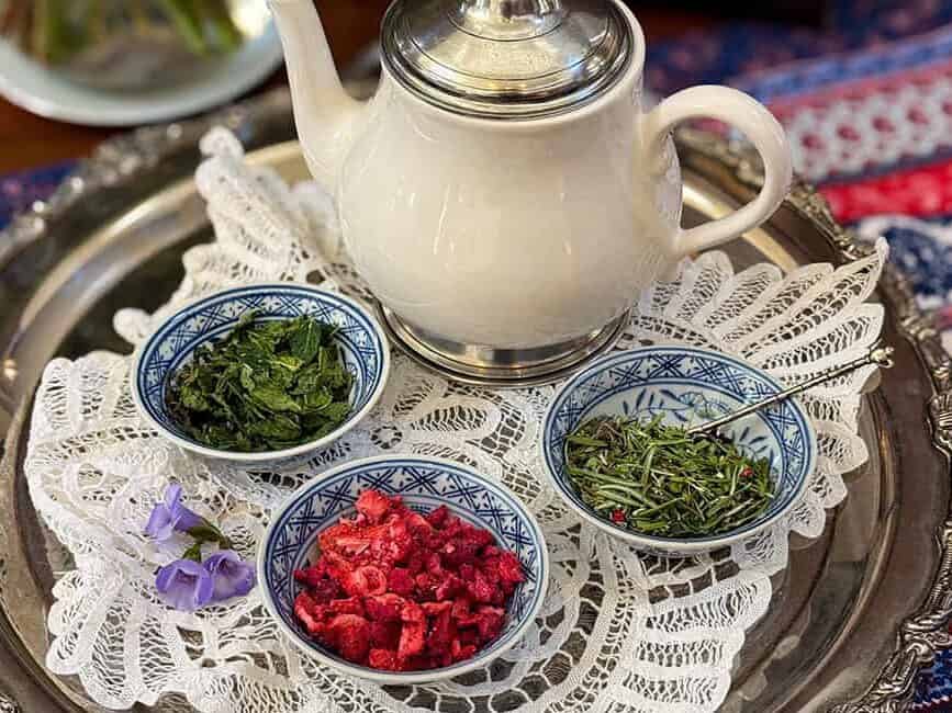 A white teapot and three small bowls containing herbs and dried berries-perfect for a Liberty Tea recipe-are arranged on a silver tray with a lace doily atop a patterned tablecloth.