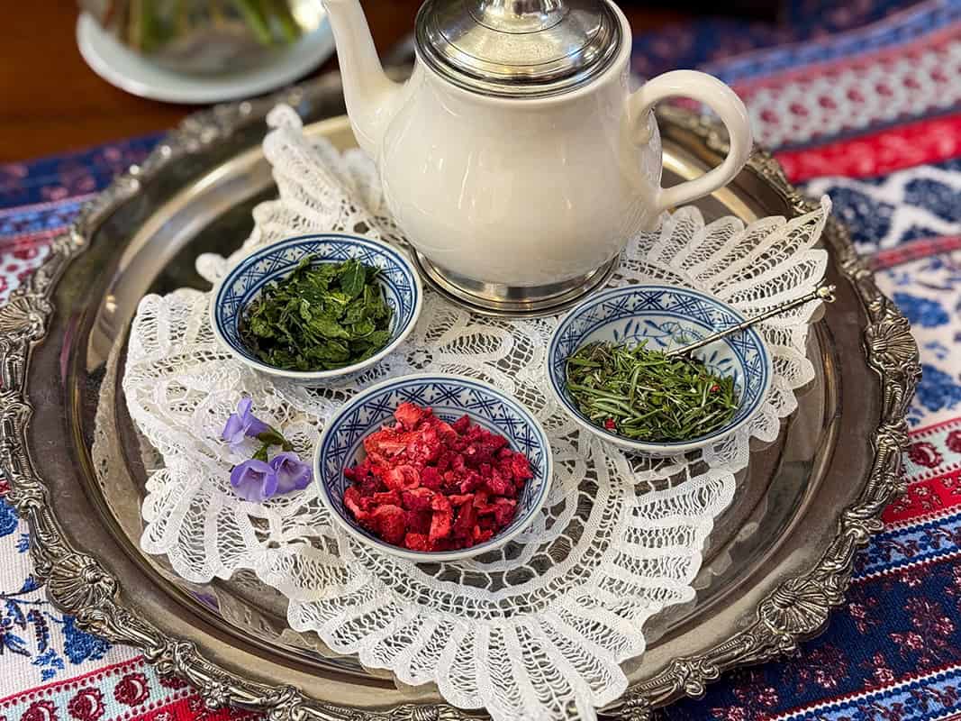A white teapot and three small bowls containing herbs and dried berries-perfect for a Liberty Tea recipe-are arranged on a silver tray with a lace doily atop a patterned tablecloth.
