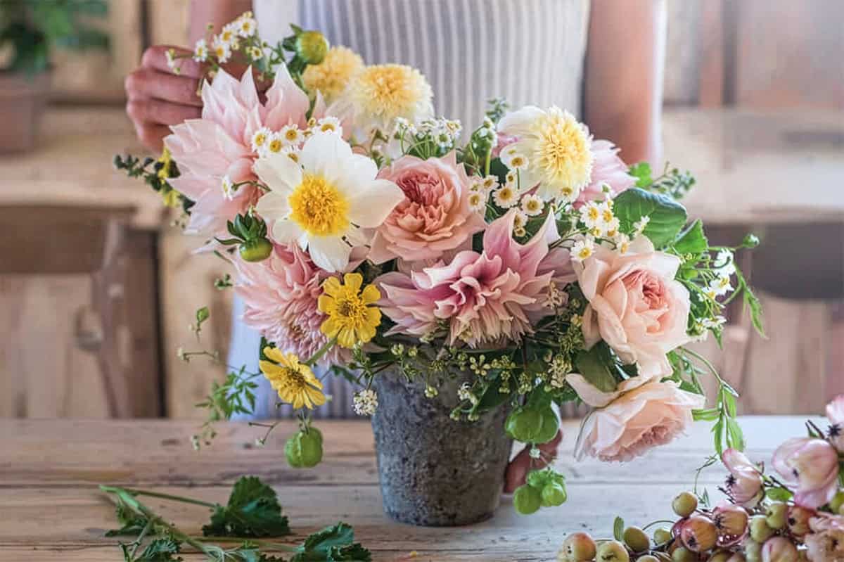 On a June afternoon tea, a person arranges a bouquet of pink, white, and yellow flowers with greenery in a rustic vase on a wooden table.