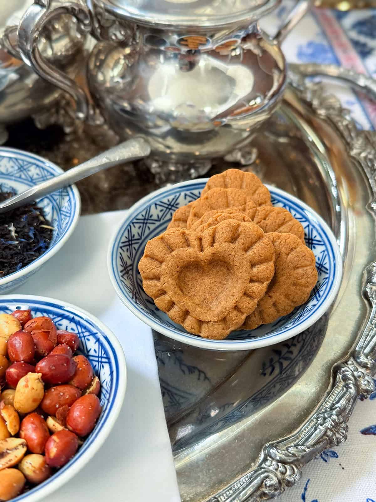 A Monticello blue and white finger bowl filled with heart-shaped gingerbread cookies sits on a silver tray next to dishes of nuts and tea, with a silver teapot in the background, evoking the charm of Colonial Williamsburg and an authentic 1796 recipe.