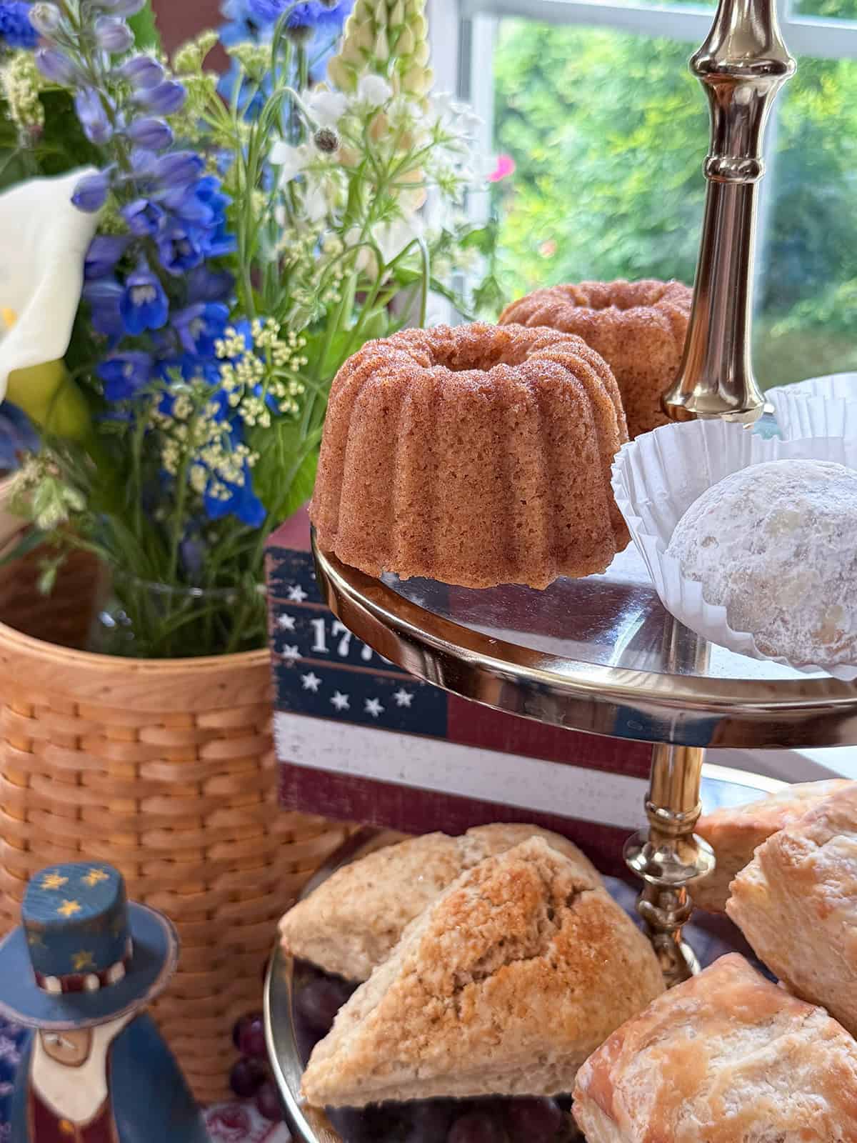 A tiered tray with mini bundt cakes, powdered sugar cookies, and scones is displayed next to a basket of flowers and revolutionary tea amid patriotic decor.