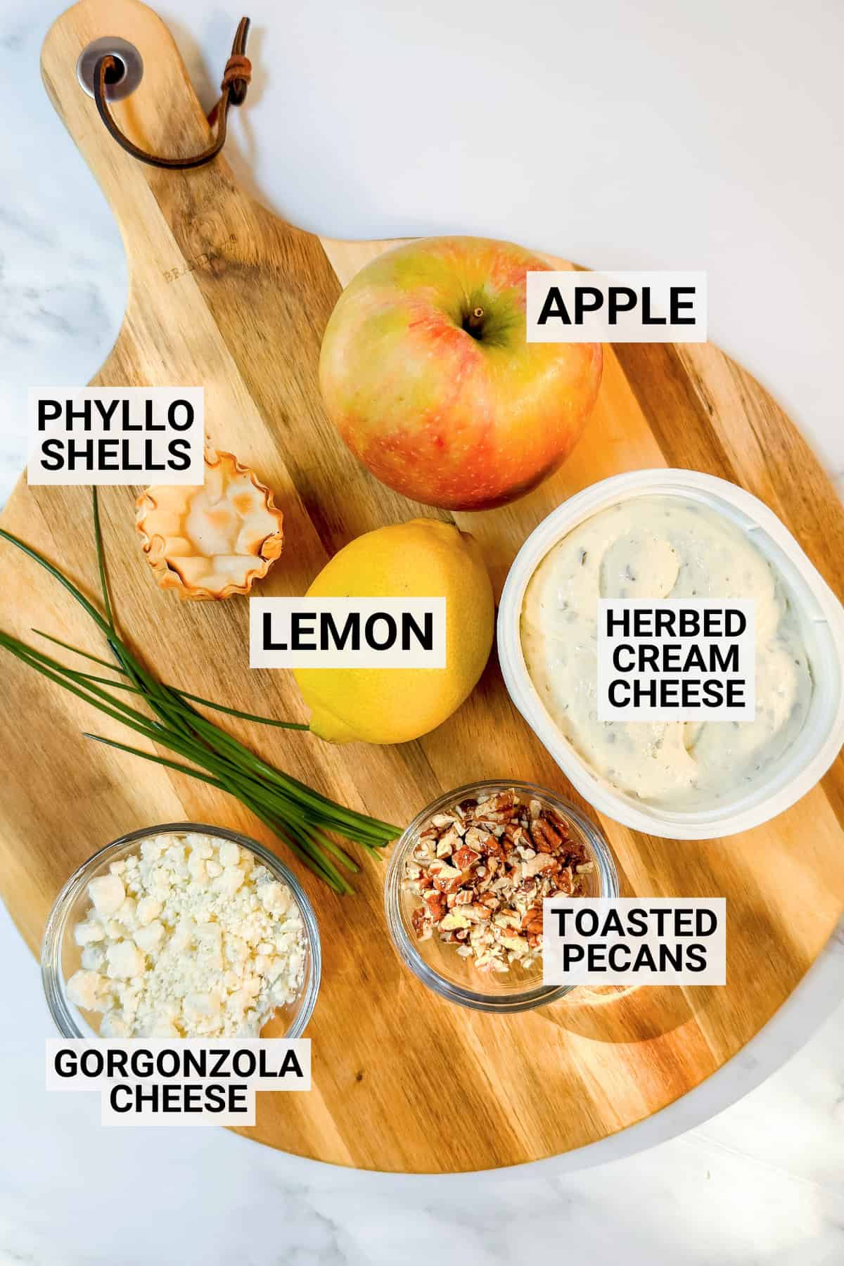 A wooden board displays apple, phyllo shells, lemon, herbed cream cheese, chives, gorgonzola cheese, and toasted pecans-perfect for assembling flavorful Apple Tartlets or savory Gorgonzola Phyllo bites. Each ingredient is labeled.