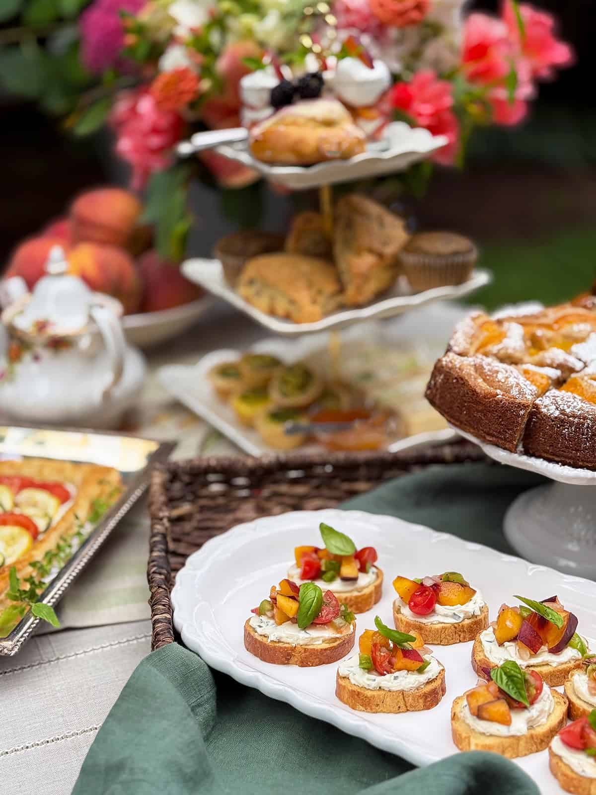 A table set with assorted pastries, peach-topped crostini, tarts, scones, and a floral arrangement in the background.