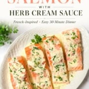 A white plate with three pieces of baked salmon with cream sauce, topped with creamy herb sauce and garnished with parsley, set on a white surface with a sprig of parsley nearby.