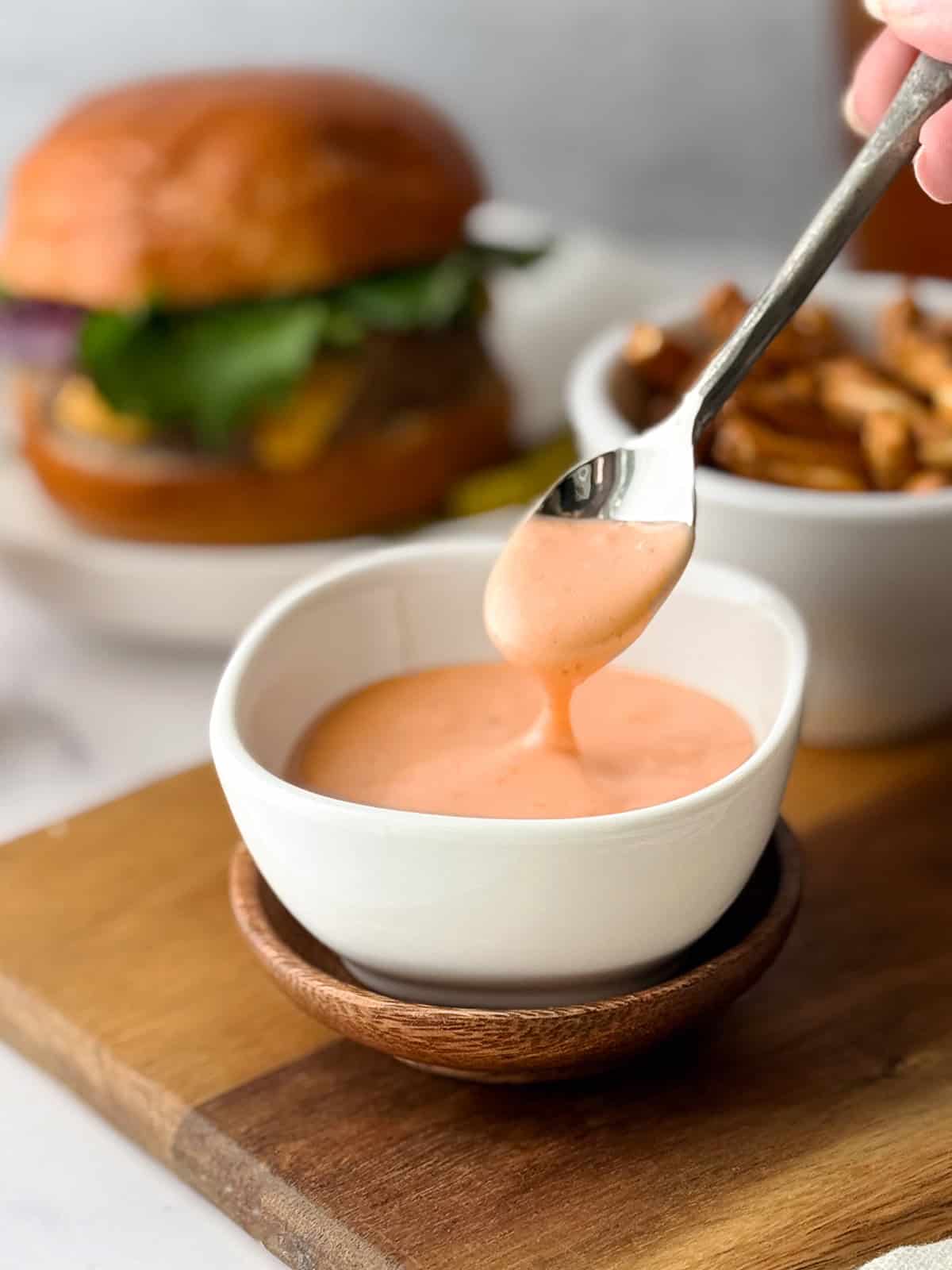 A spoon drizzling homemade burger sauce into a small white bowl, with a burger and fries blurred in the background.