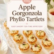 A platter of Gorgonzola tartlets and apple phyllo tartlets is arranged on a lace-trimmed tray beside tea cups and pink flowers, with a sign describing them as savory tea-time appetizers.