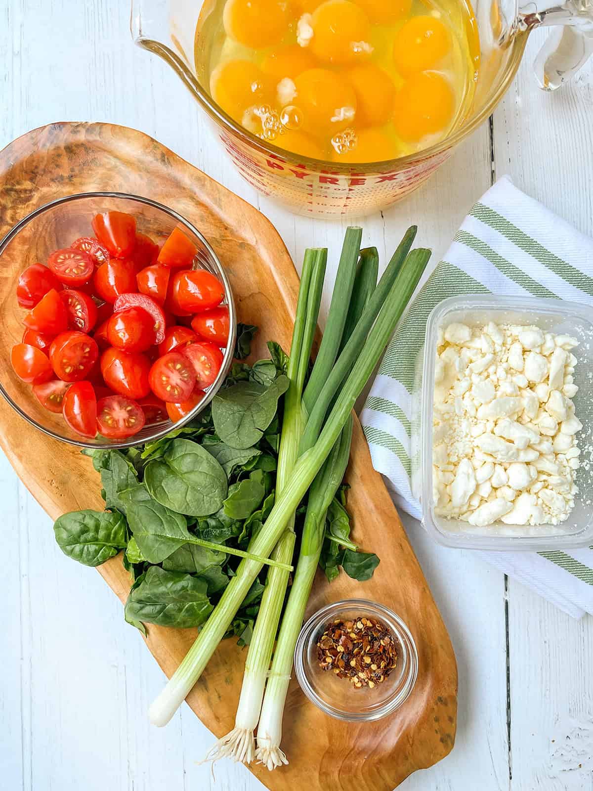 A wooden board with spinach, green onions, cherry tomatoes, and chili flakes next to a container of feta and a measuring cup of cracked eggs on a white surface-perfect for creating quick and easy recipes.