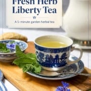 A blue and white teacup filled with Liberty Tea sits on a saucer next to fresh herbs, a teapot, a cookie, and blue flowers on the table.