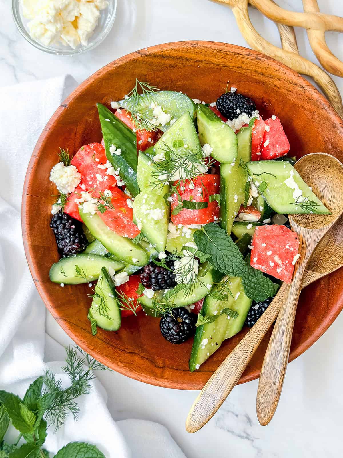 A wooden bowl of salad with cucumber, watermelon, blackberries, feta cheese, fresh mint, and dill offers a refreshing twist on quick and easy recipes, served with wooden salad servers on the side.