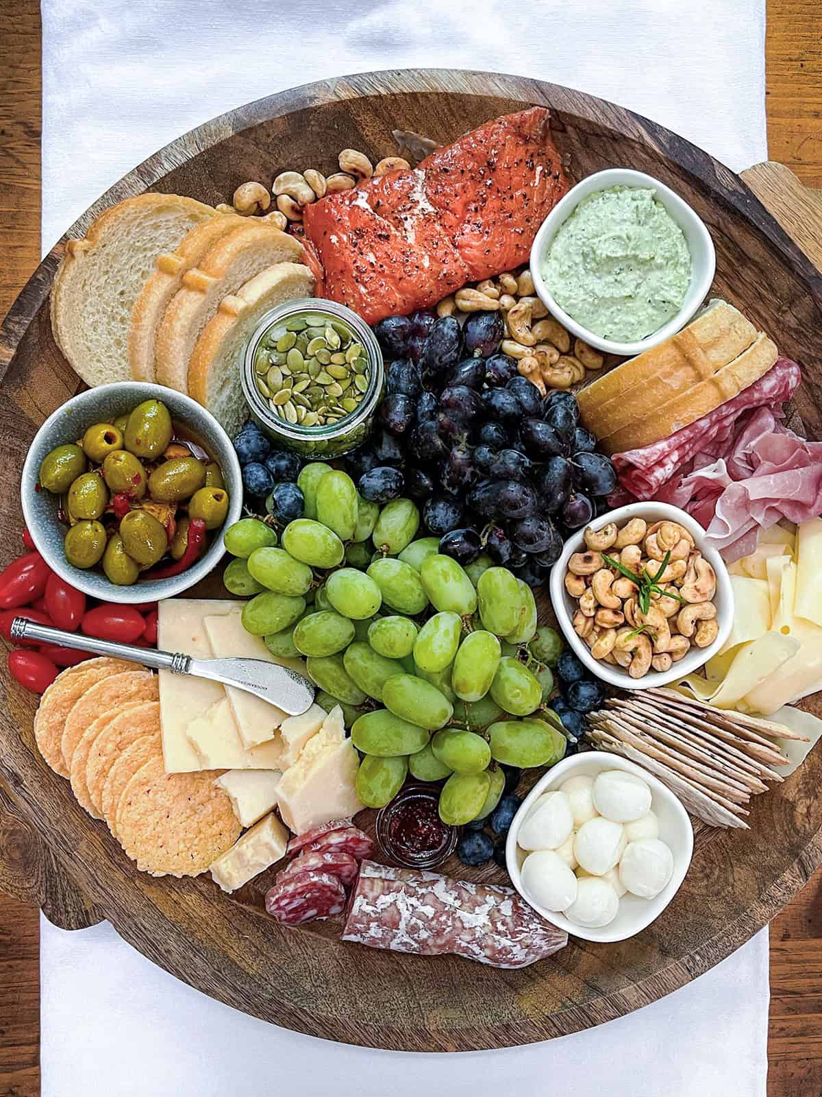 A wooden platter with bread, cheeses, grapes, olives, cured meats, smoked salmon, nuts, crackers, mozzarella balls, dip, and pumpkin seeds arranged in sections.