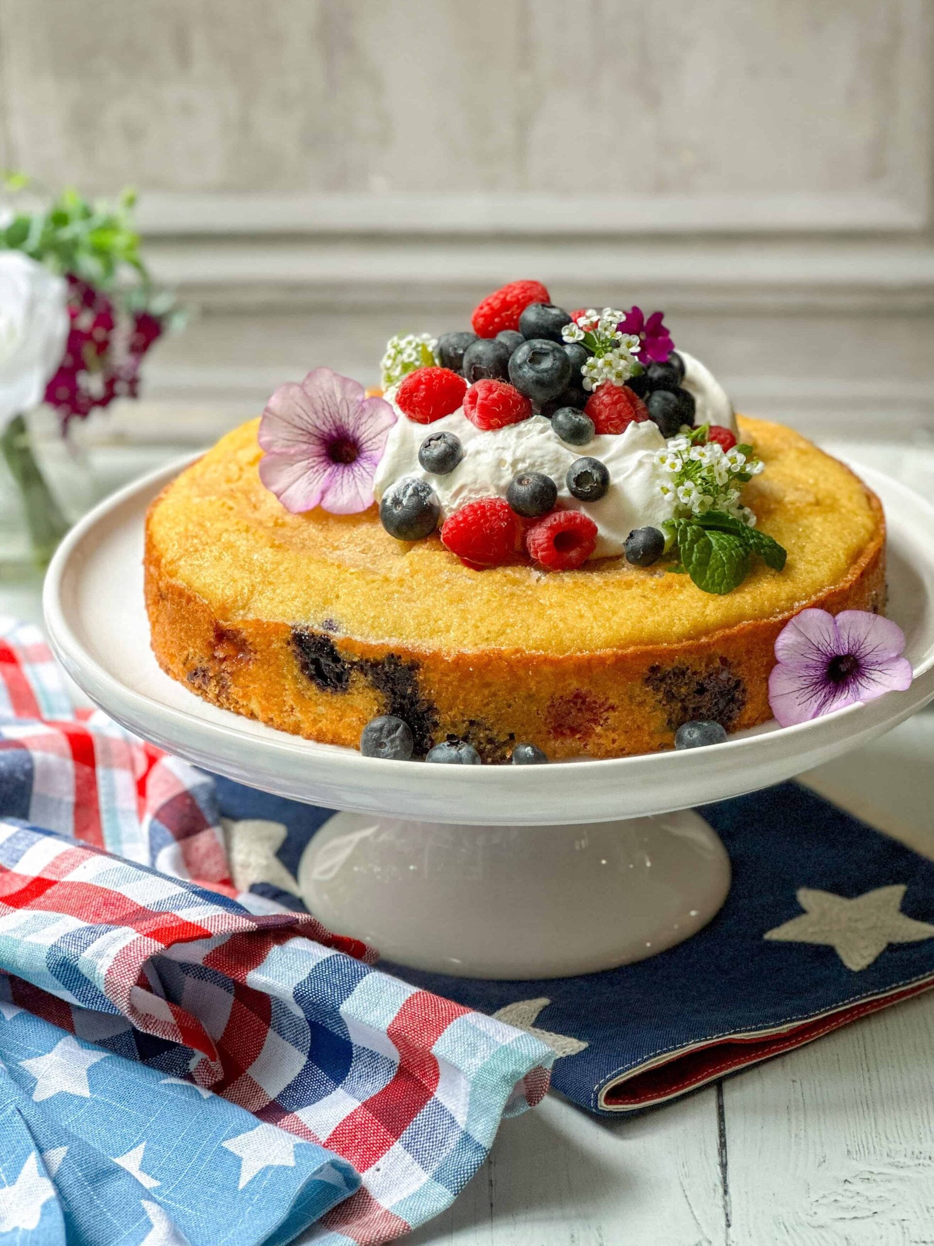 A round berry cake topped with whipped cream, blueberries, raspberries, and edible flowers sits on a white cake stand next to a star-patterned cloth.
