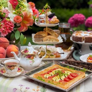A table set for afternoon tea with floral china, a teapot of lavender tea, fresh fruit, pastries, and a vegetable tart, surrounded by flowers and greenery.
