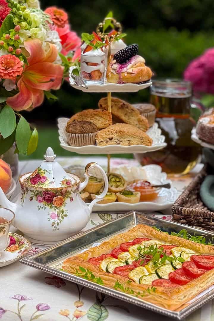A table set for afternoon tea with floral china, a teapot of lavender tea, fresh fruit, pastries, and a vegetable tart, surrounded by flowers and greenery.