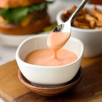 A spoon drizzles creamy orange homemade burger sauce into a small white bowl, with fries and a burger blurred in the background.