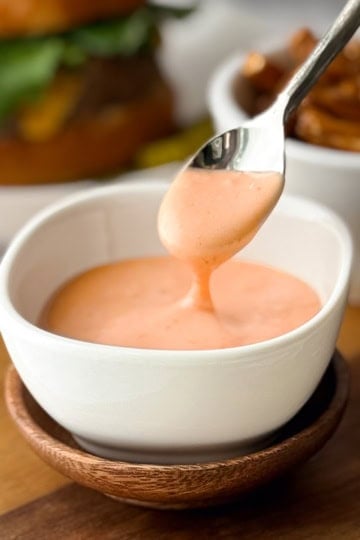 A spoon drizzles creamy orange homemade burger sauce into a small white bowl, with fries and a burger blurred in the background.