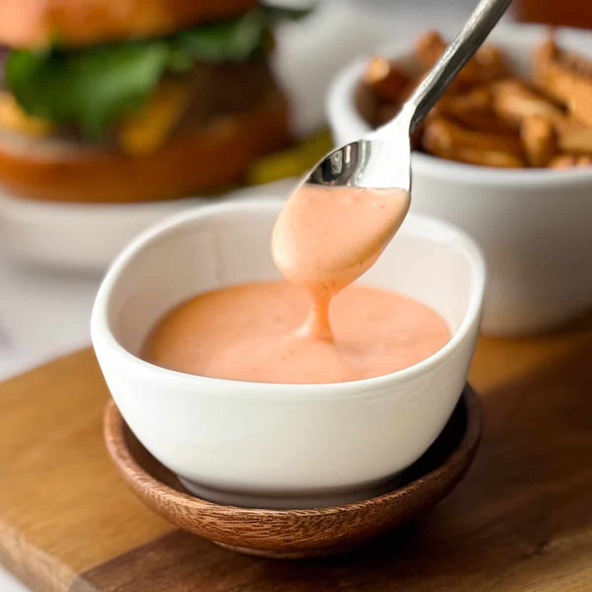 A spoon drizzles creamy orange homemade burger sauce into a small white bowl, with fries and a burger blurred in the background.