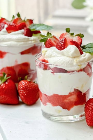 Two glass cups filled with layers of whipped cream and strawberries sit on a counter, showcasing one of our favorite quick and easy recipes. Each cup is garnished with fresh strawberries and mint leaves, while whole strawberries rest nearby.