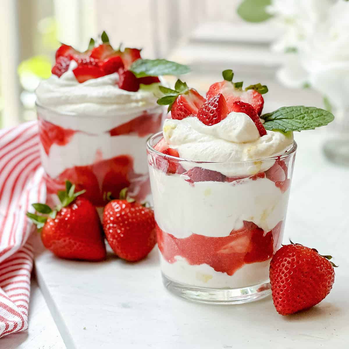 Two glass cups filled with layers of whipped cream and strawberries sit on a counter, showcasing one of our favorite quick and easy recipes. Each cup is garnished with fresh strawberries and mint leaves, while whole strawberries rest nearby.