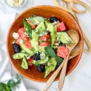A wooden bowl of sliced cucumbers, watermelon, blackberries, feta cheese, and herbs sits on a white surface with two wooden spoons beside it-a fresh and vibrant idea for those seeking quick and easy recipes.