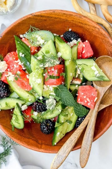A wooden bowl of sliced cucumbers, watermelon, blackberries, feta cheese, and herbs sits on a white surface with two wooden spoons beside it—a fresh and vibrant idea for those seeking quick and easy recipes.