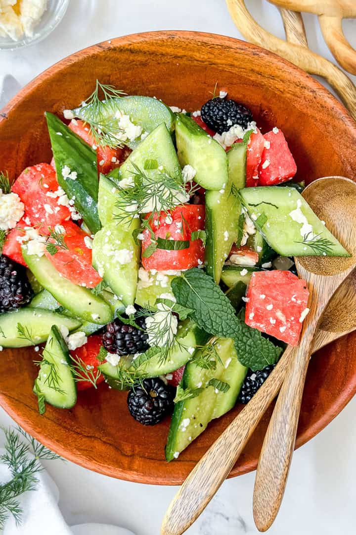 A wooden bowl of sliced cucumbers, watermelon, blackberries, feta cheese, and herbs sits on a white surface with two wooden spoons beside it-a fresh and vibrant idea for those seeking quick and easy recipes.