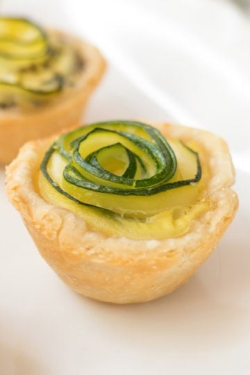 Mini tart with a golden crust, filled with custard and crumbled feta, and topped with thinly sliced zucchini arranged in a rosette shape, resembling elegant zucchini quiches, placed on a white plate.