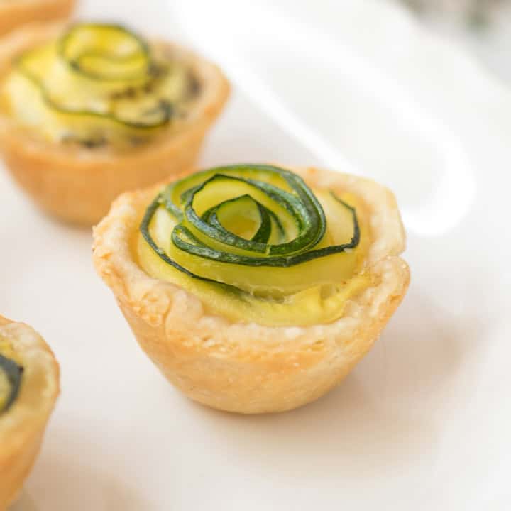Mini tart with a golden crust, filled with custard and crumbled feta, and topped with thinly sliced zucchini arranged in a rosette shape, resembling elegant zucchini quiches, placed on a white plate.