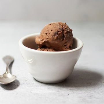 A scoop of Easy Chocolate Ice Cream in a white ceramic bowl sits next to a silver spoon on a light-colored surface.