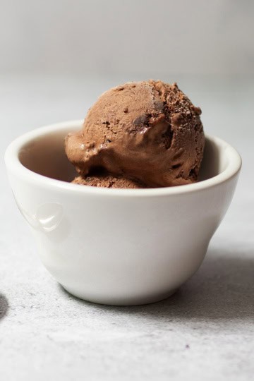 A scoop of Easy Chocolate Ice Cream in a white ceramic bowl sits next to a silver spoon on a light-colored surface.
