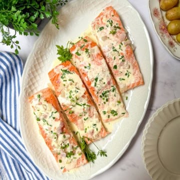 Four baked salmon fillets topped with a rich herb cream sauce and chopped herbs are arranged on a white oval platter. This elegant baked salmon recipe is complemented by a striped napkin, green herb plant, and small potatoes nearby.