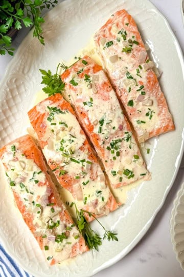 Four baked salmon fillets topped with a rich herb cream sauce and chopped herbs are arranged on a white oval platter. This elegant baked salmon recipe is complemented by a striped napkin, green herb plant, and small potatoes nearby.