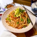 A plate of Cold Peanut Sesame Noodles with sliced vegetables and herbs, garnished with cucumber and mint, with chopsticks resting on the plate.
