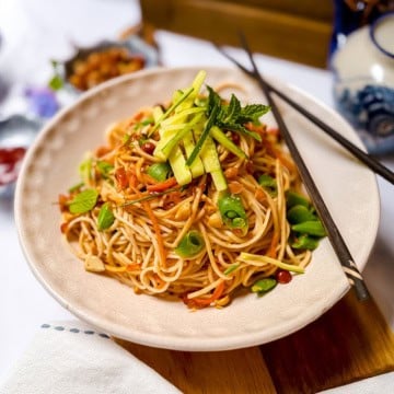 A plate of Cold Peanut Sesame Noodles with sliced vegetables and herbs, garnished with cucumber and mint, with chopsticks resting on the plate.