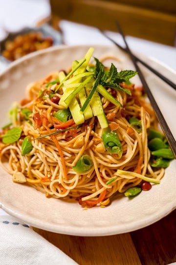 A plate of Cold Peanut Sesame Noodles with sliced vegetables and herbs, garnished with cucumber and mint, with chopsticks resting on the plate.